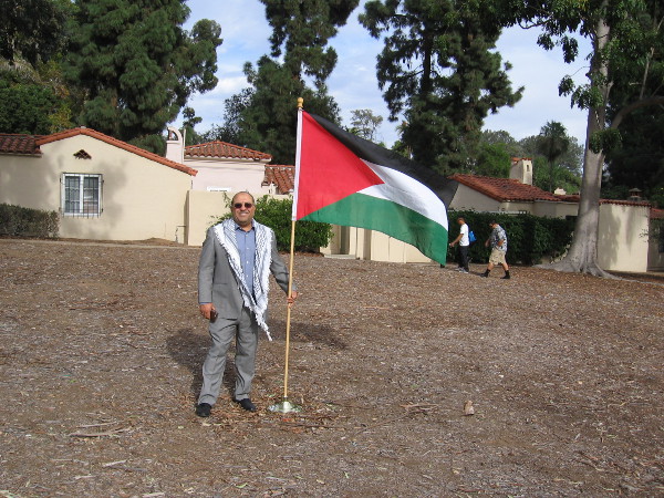A gentleman from the House of Palestine stands near a flag where a new cottage will be built that showcases Palestinian culture.
