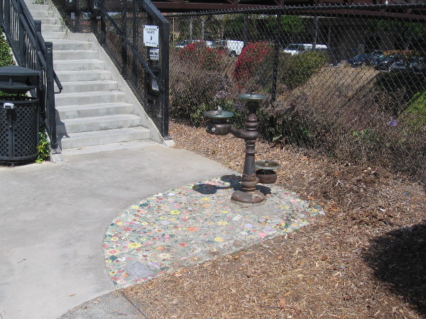 A water fountain near steps to the Dahlia Drive pedestrian bridge that spans the train tracks. The fountain stands above colorful mosaics.